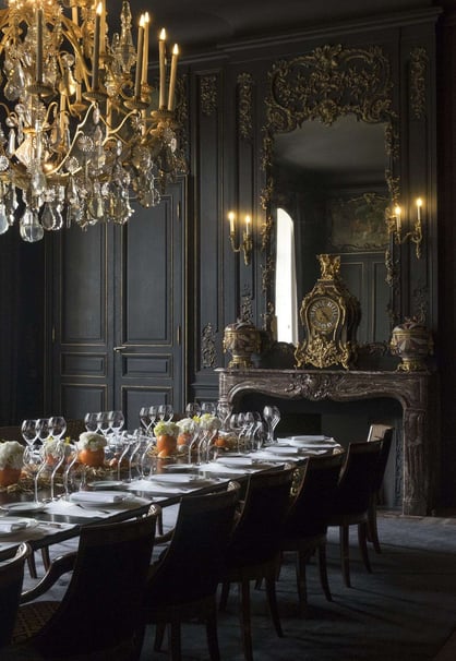 gradn dining room with ornate black wood wall panelling  and long dining table with rows of chairs. antique gold mirror and chandelier and candle scones bring warmth to the room 